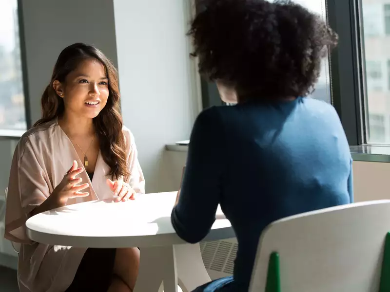 Two women wearing business attire having a discussion at a white table in front of a window.